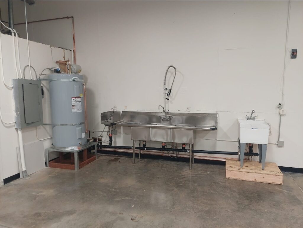 Commercial kitchen area featuring a large stainless steel sink, a hot water heater, and a smaller utility sink, highlighting food preparation facilities.