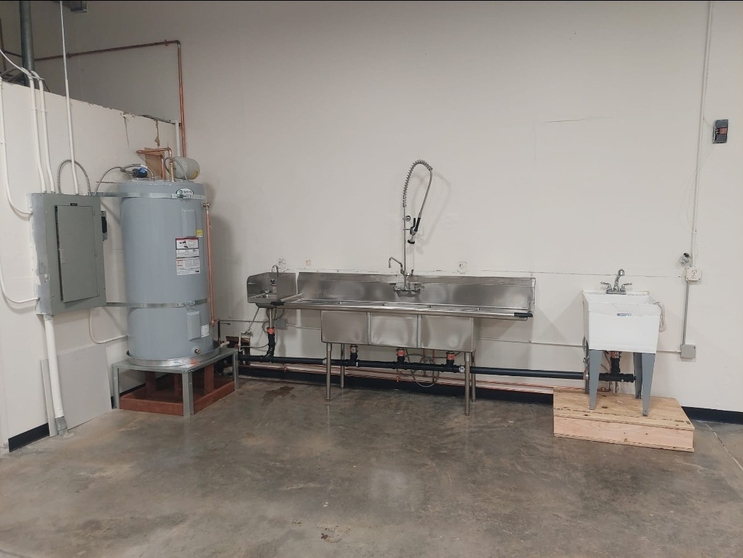Commercial kitchen area featuring a large stainless steel sink, a hot water heater, and a smaller utility sink, highlighting food preparation facilities.