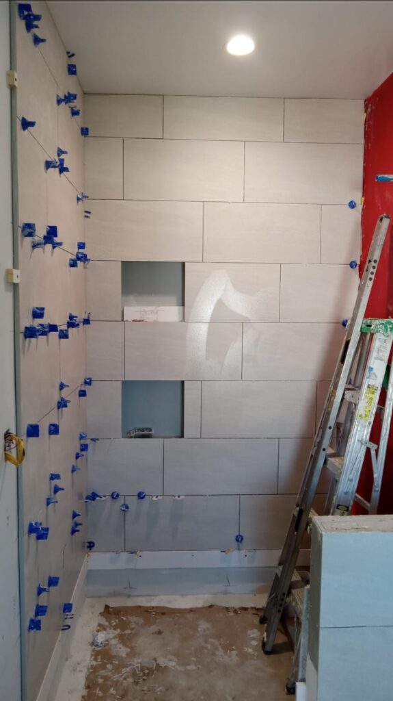 Light-colored tiled wall in progress, featuring a recessed section. Blue spacers are used for precise alignment. A ladder and tools are visible, indicating ongoing renovation.