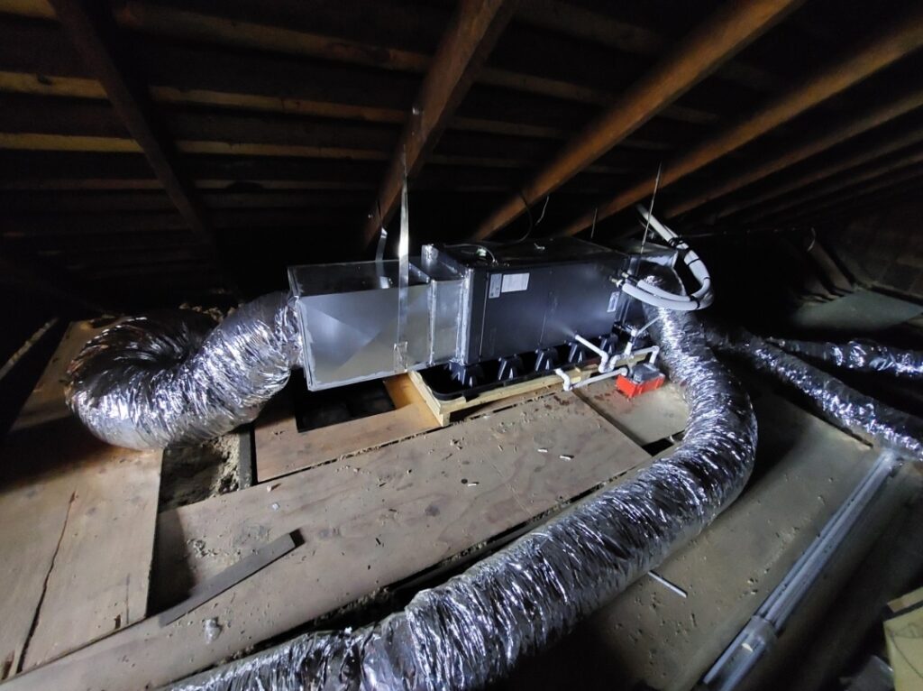 HVAC system in an attic, featuring silver ductwork and a black air handler on wooden beams, illustrating home ventilation setup.
