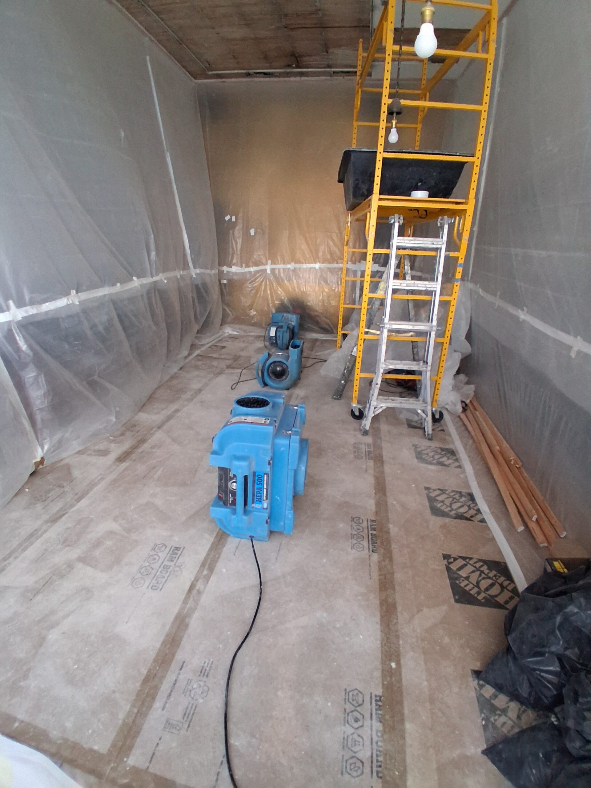 Construction site featuring plastic sheeting enclosing the area, two blue air movers, a ladder, and a scaffold, indicating ongoing renovation work.
