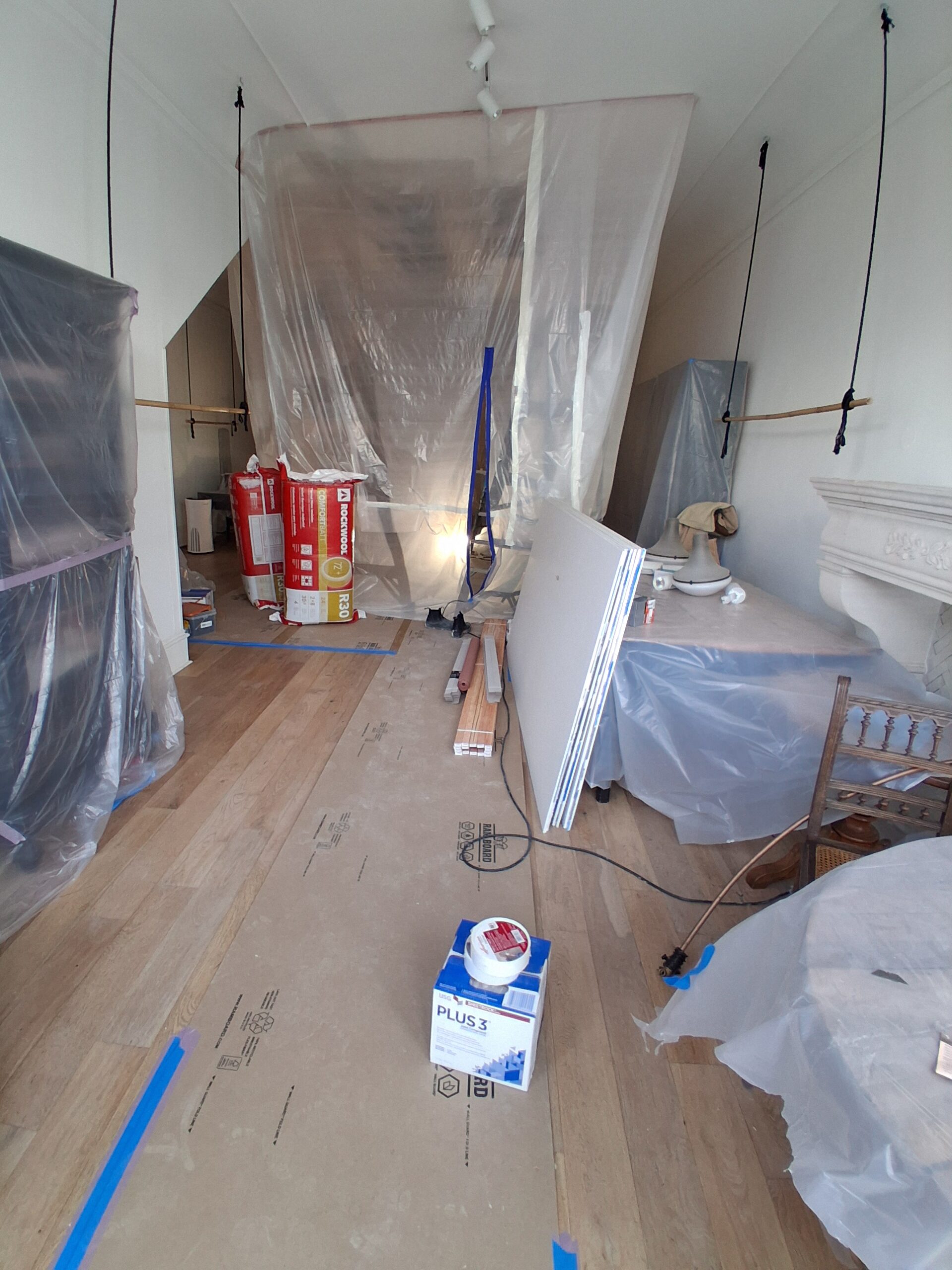 Renovation workspace with plastic sheeting, construction materials, and tools scattered across the wooden floor, indicating ongoing remodeling.