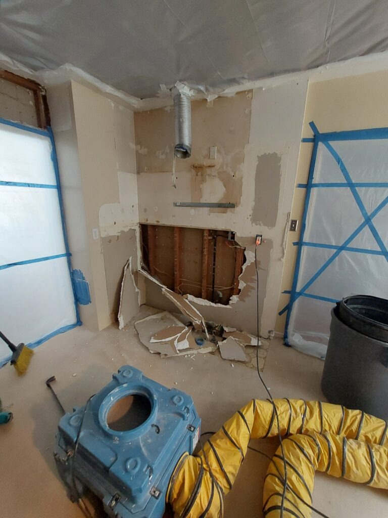 Partially demolished wall in a room undergoing renovation, featuring exposed studs, a blue machine, and yellow ducting for airflow.