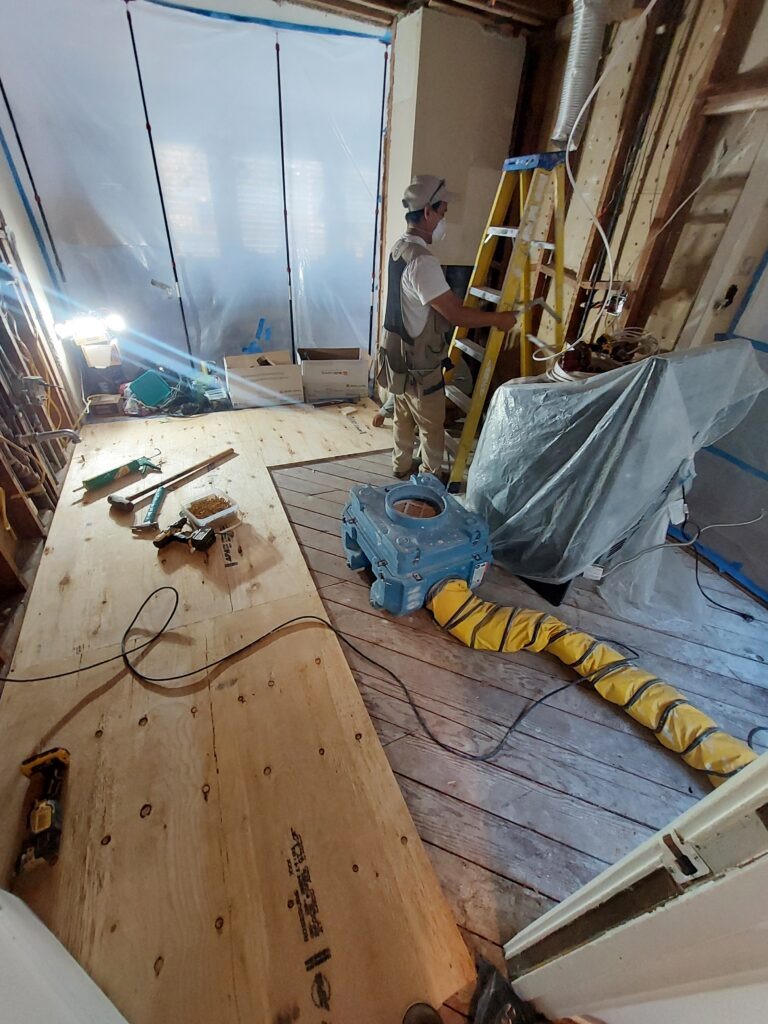 A construction worker stands on a ladder, preparing to work in a partially renovated room enclosed with plastic sheeting. Tools and equipment are scattered around.