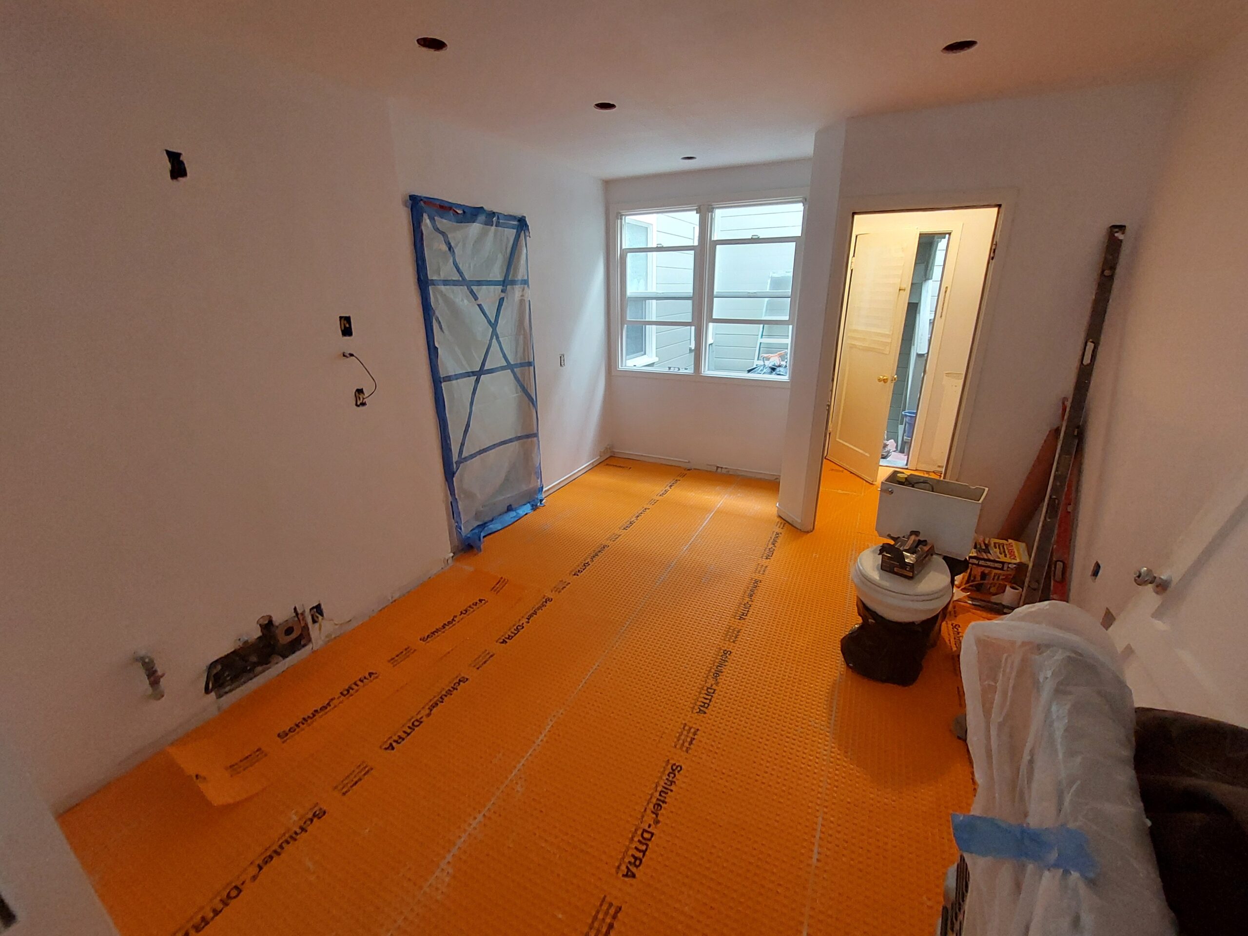 Renovated room featuring new orange flooring, exposed walls, and a toilet on a garbage bag. Natural light enters through a nearby window.