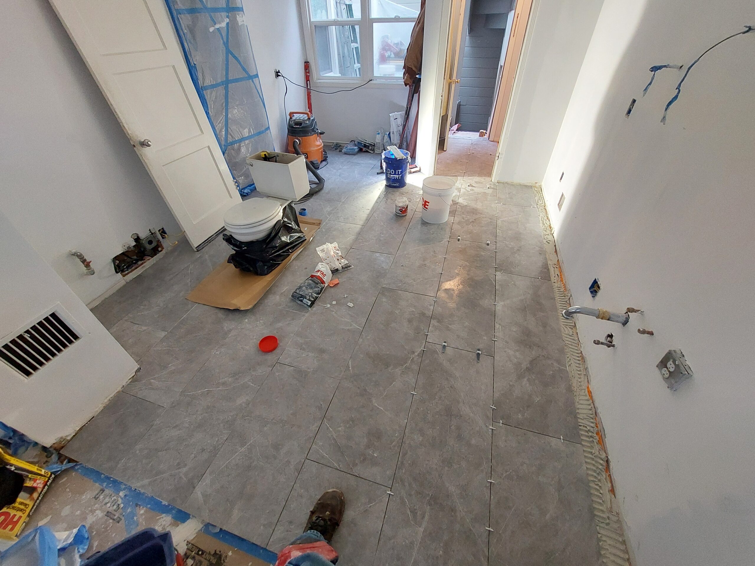 Floor tiling in a partially renovated room, featuring a newly laid gray tile floor, a toilet on cardboard, and construction debris scattered around.
