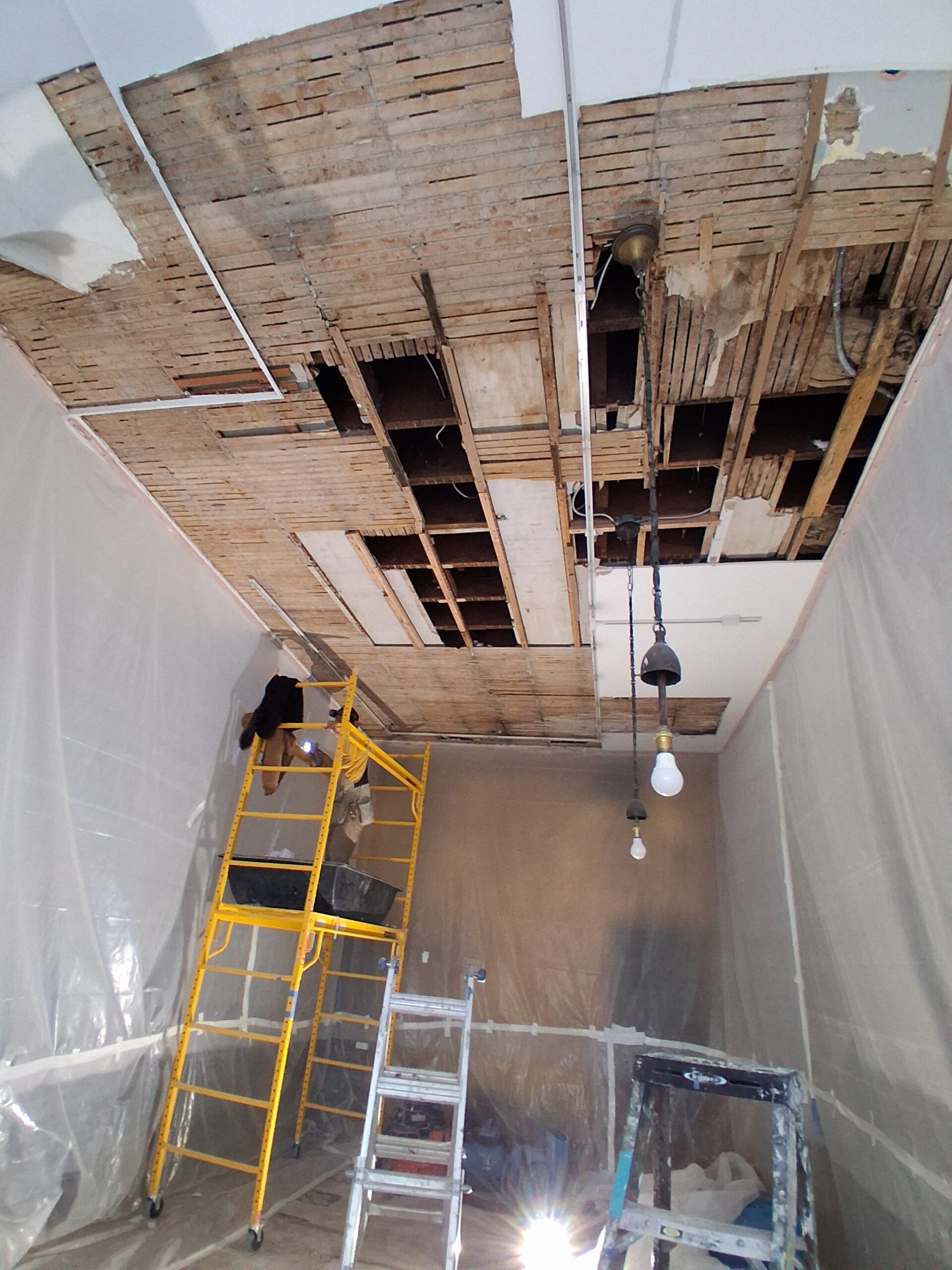 Ceiling undergoing renovation shows exposed beams and wiring. Two ladders are present, with a worker on a yellow scaffold preparing for repairs.