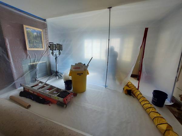 A construction site enclosed in plastic sheeting, featuring work lights, a ladder, a yellow waste bin, and ventilation ducting.