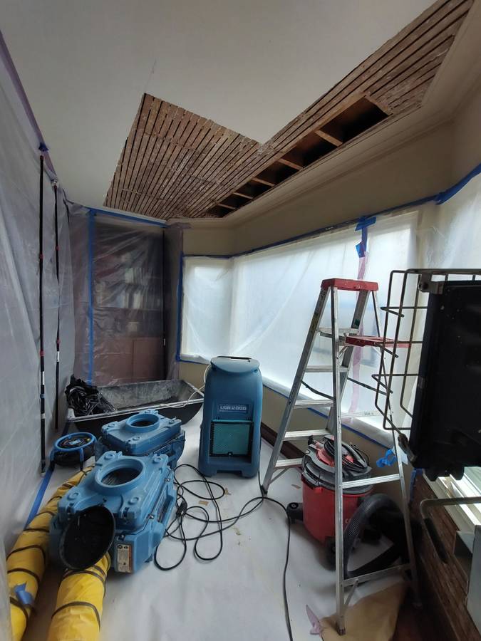 Renovation scene showing exposed ceiling beams in a room under construction. Tools and equipment are scattered on a protected floor.