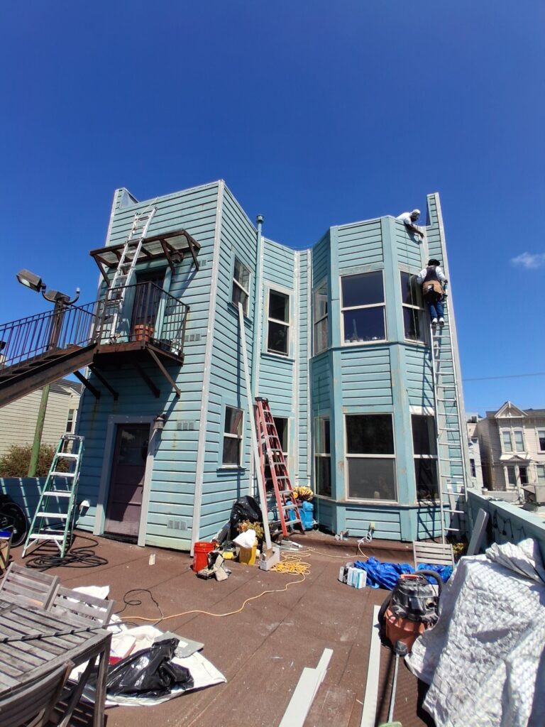 A light blue two-story house under renovation, featuring ladders and construction equipment scattered around, with one worker on a ladder against the wall under a clear blue sky.